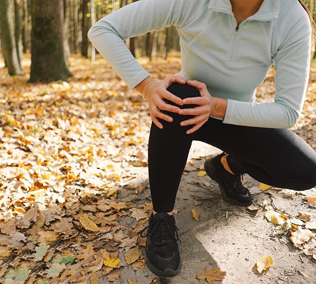 A woman in activewear surrounded by woods and fallen leaves holds her right knee with both hands