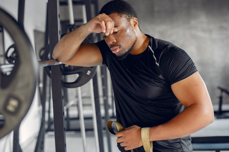 A young Black adult man rests his elbow on a while sitting at a bench press machine with his hand on his forehead