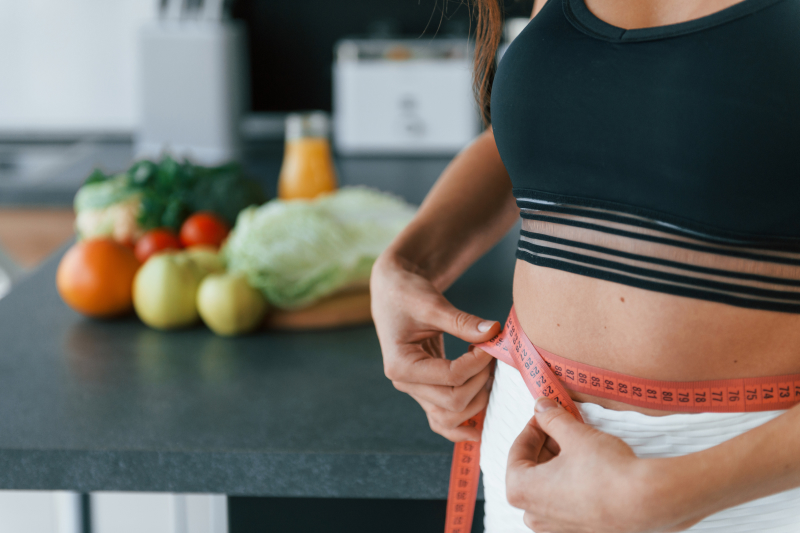 A woman holds a sewing tape measure to her waist in the foreground and vegetables in the background on a kitchen island
