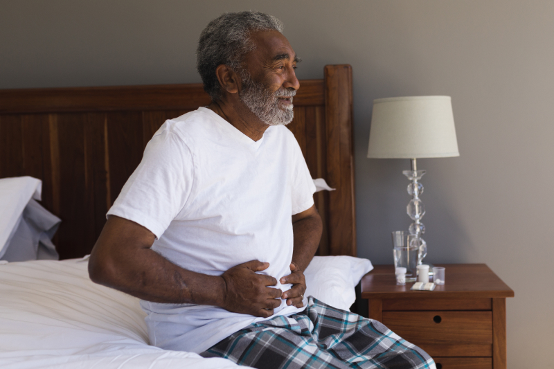 Older adult man with a grimaced face holds his stomach in a bedroom