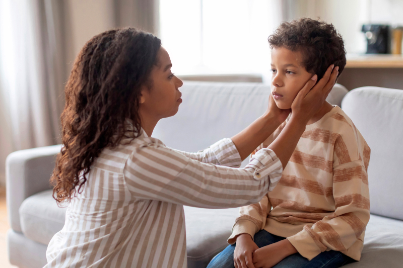 Concerned Black mother tenderly holding her son’s face at home