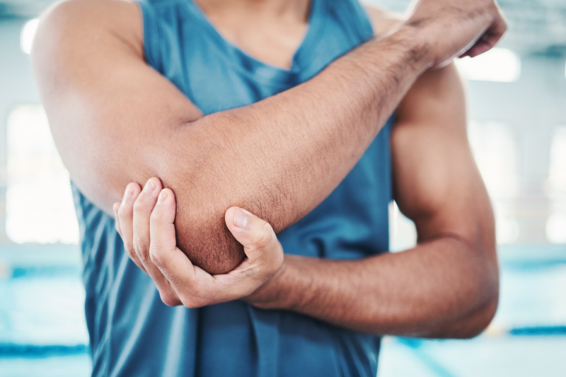 Man in blue tank top holding right elbow with left hand with a swimming pool in the background
