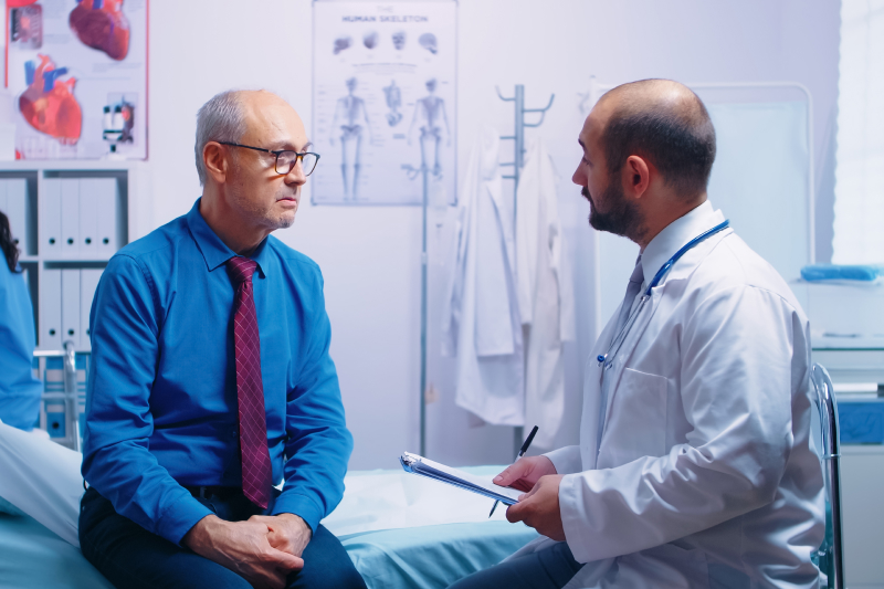 Older adult man sits on examination table in medical appointment room and speaks to a medical provider with a clipboard