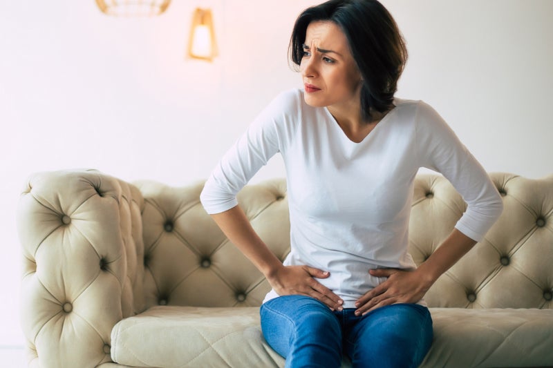 Young woman sits on a sofa and touches her lower stomach and grimaces in pain