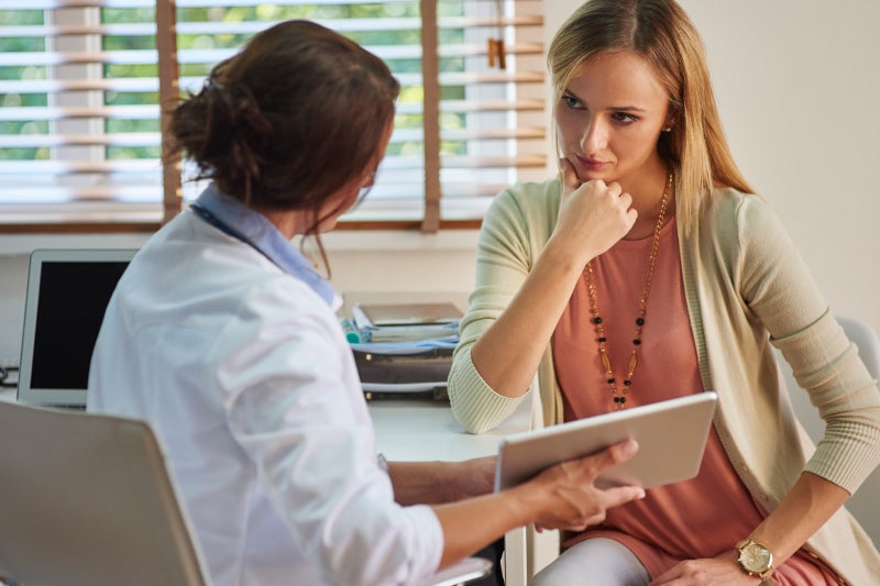 A woman in casual attire consults with a doctor in an office; the doctor holds a tablet while the woman listens intently