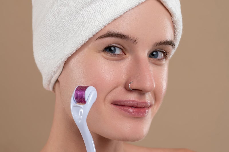 Smiling woman with a towel wrapped on her head uses a dermaroller on her cheek