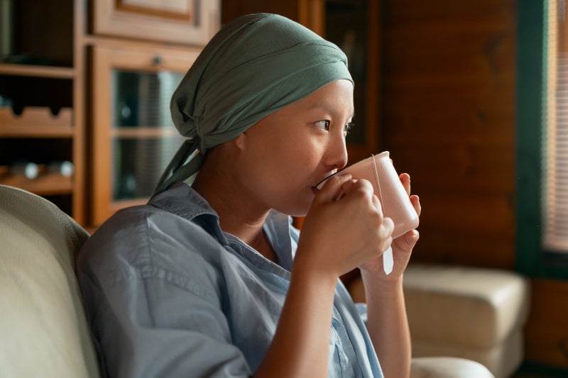 A person in a light blue shirt and green headscarf sits on a couch, sipping from a mug; the room is warmly lit with wooden walls and a window