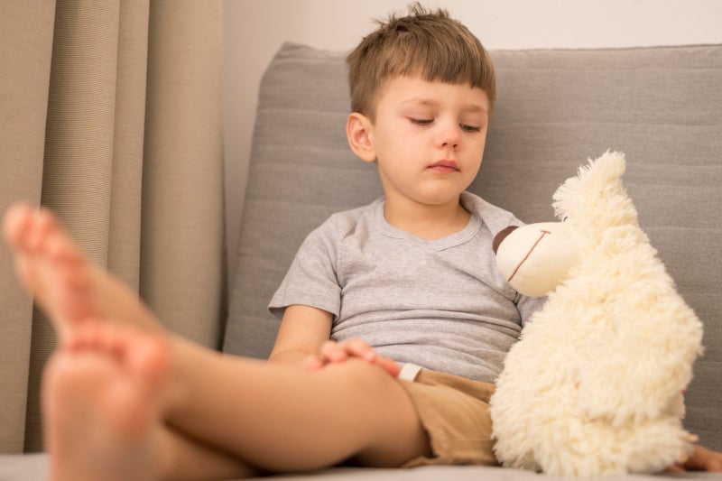 Young boy sitting on a couch holding a white stuffed animal, looking sad with his legs stretched out in front of him