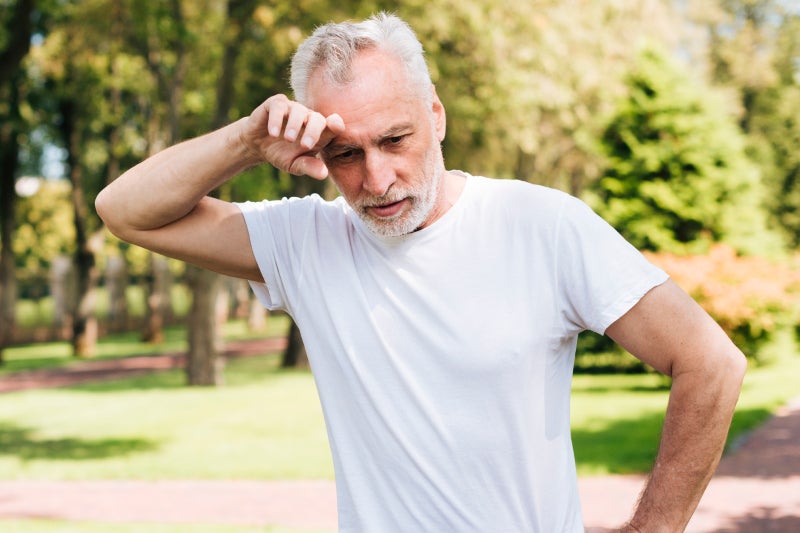 An older adult man stands in a park, with lush trees and grass in the background, appearing relaxed and content