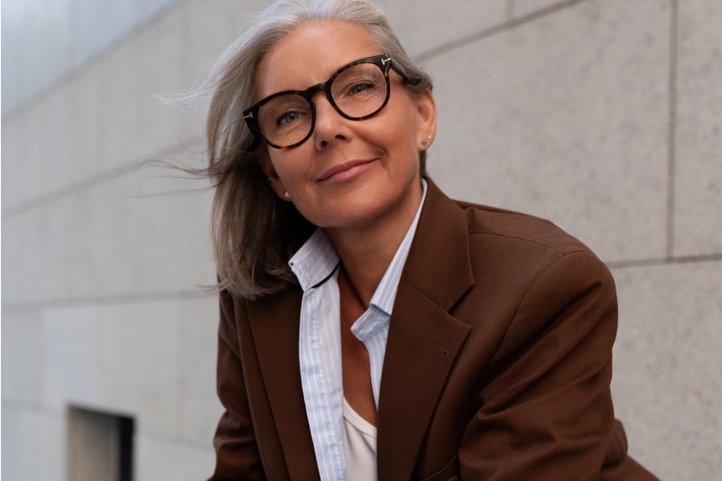 A woman wearing glasses and a brown jacket leans against a wall while looking relaxed and smiling
