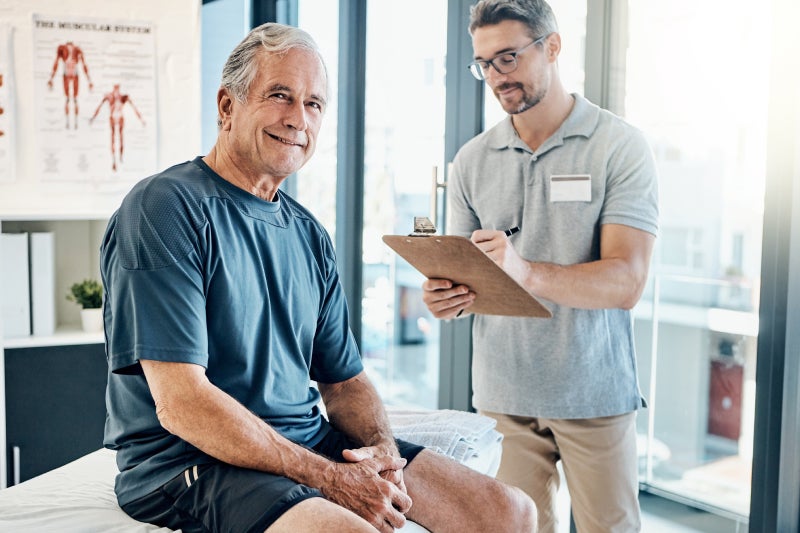 Smiling older man sits on a clinic exam table while a healthcare provider with a clipboard consults him; anatomy charts and windows are behind them