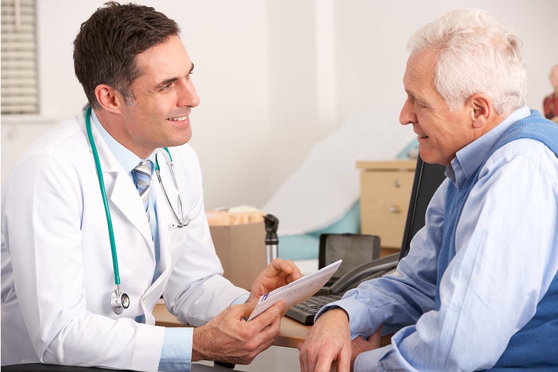 Doctor in a white coat smiles while discussing paperwork with an elderly male patient in a wheelchair during a friendly medical consultation