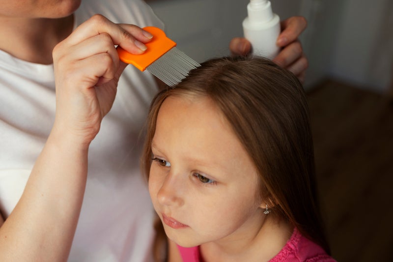 An adult carefully checks a young girl’s brown hair with an orange nit comb, holding a bottle nearby; the child looks calm and focused