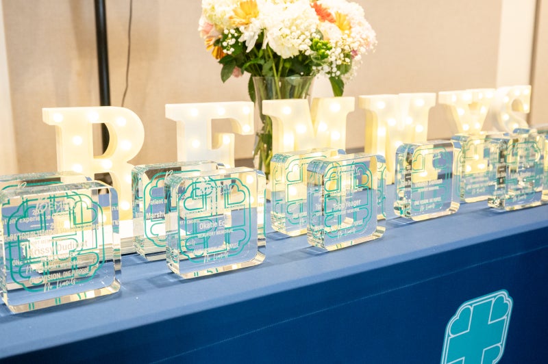 Glass awards sit in front of light up letters spelling BEMMYS on a table draped with a blue tablecloth.