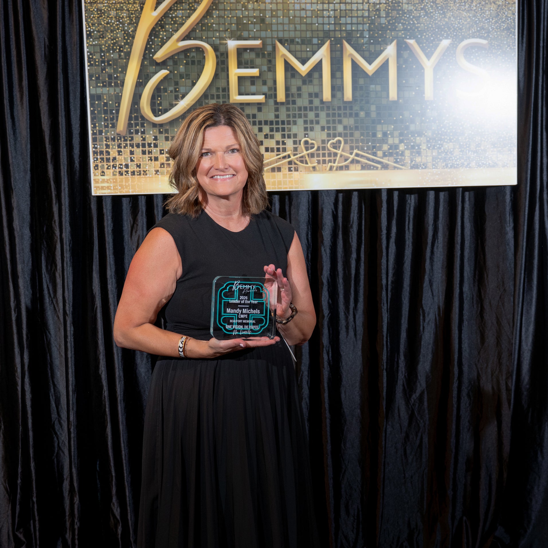 Mandy Michels holds a 2026 Bemmy Award in front of a black backdrop and hanging sign that reads BEMMYS.