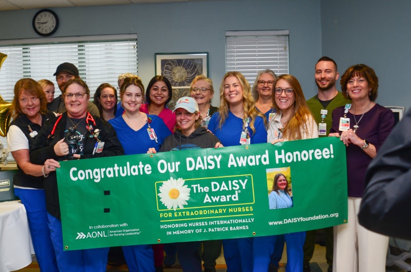 Beaufort Memorial employees join ICU nurse Candy Chappell and her family for a photo behind a DAISY Award recognition banner