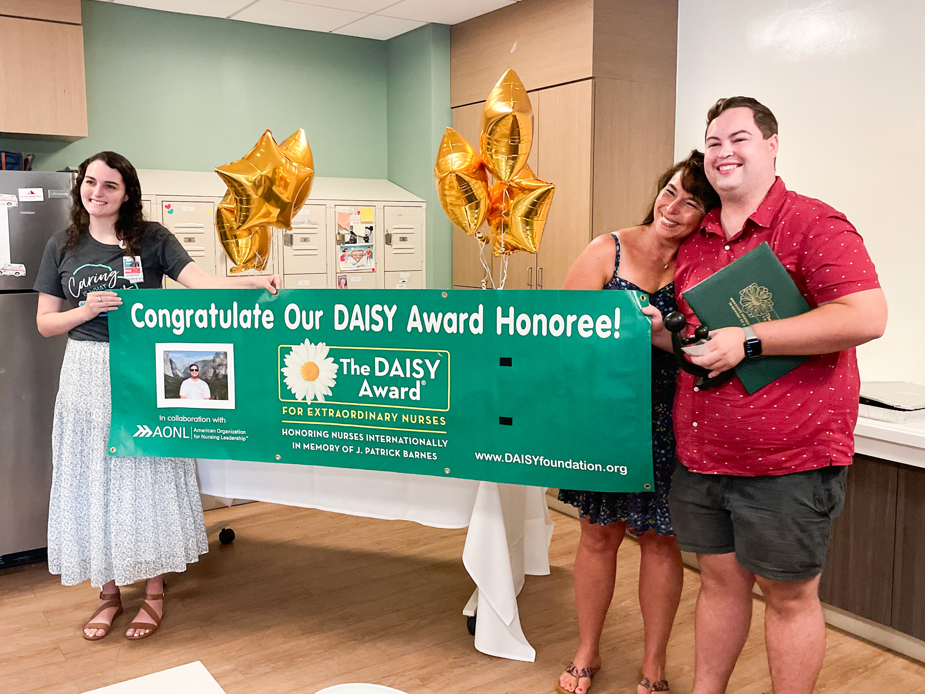 Jonathan Barrett stands behind a celebratory banner for his DAISY Award, posing with his mother and a member of the Beaufort Memorial education team 