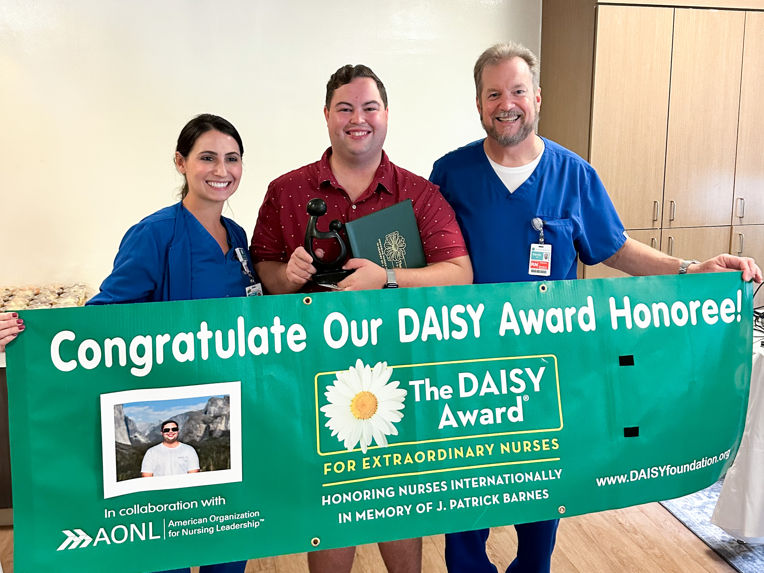 Andrea Smith, Jonathan Barrett and Kevin Kremer stand behind a banner recognizing Jonathan Barrett for receiving a DAISY Award