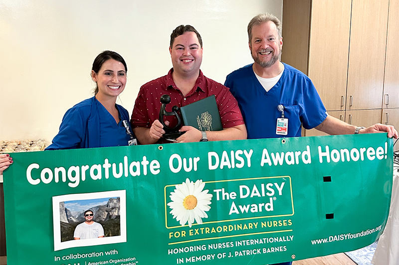 Andrea Smith (left) and Kevin Kremer (right) hold a congratulations banner for Jonathan Barrett for being named a DAISY award recipient