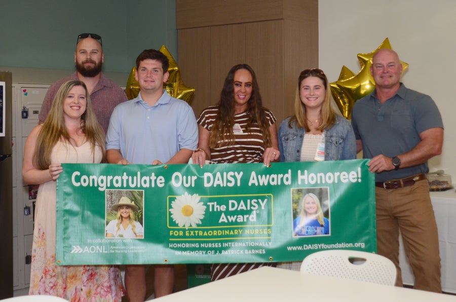 Kaitlyn Heslin, RN poses for a photo with her family and her coworker Michelle Recchia in front of a Congratulate Our DAISY Award Honoree banner.