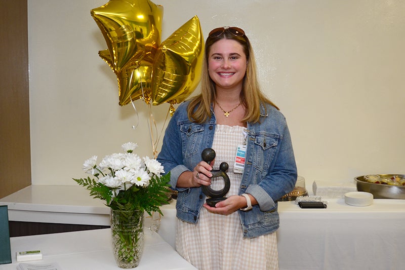 Kaitlyn Heslin, a registered nurse with the Beaufort Memorial Emergency Department, holds a DAISY award standing next to a vase of daisies on a table.