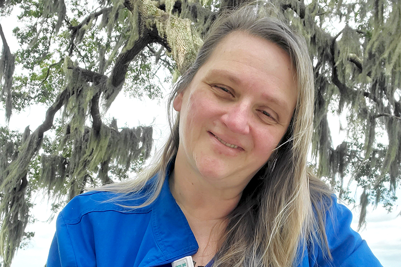 Denise Briggs, RN, a nurse with Beaufort Memorial, smiles at the camera with spanish moss covered live oaks behind her