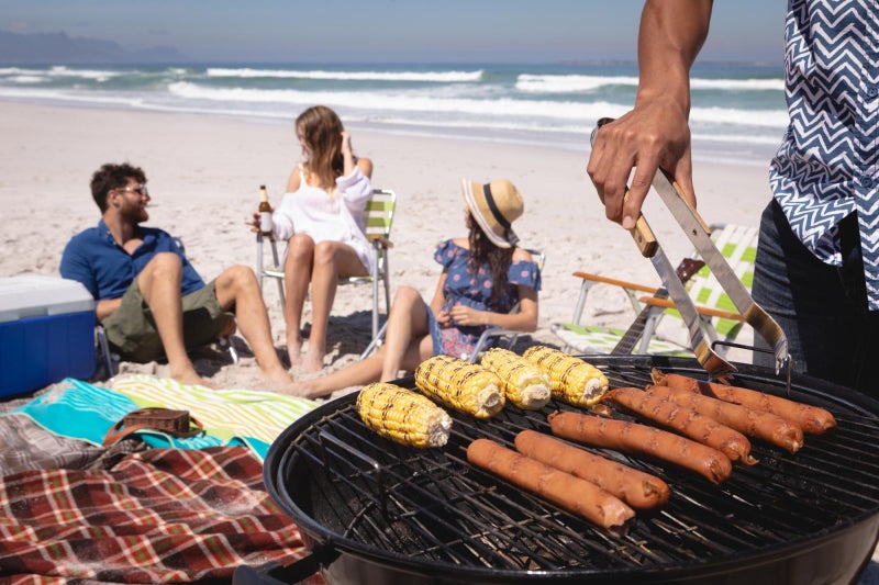 Diverse group of friends sitting on the beach with one person in the foreground grilling corn and hot dogs.