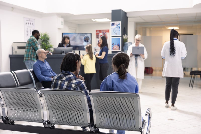 A wide angle photo of a somewhat busy emergency room waiting area with people sitting in seats and standing up in various places.