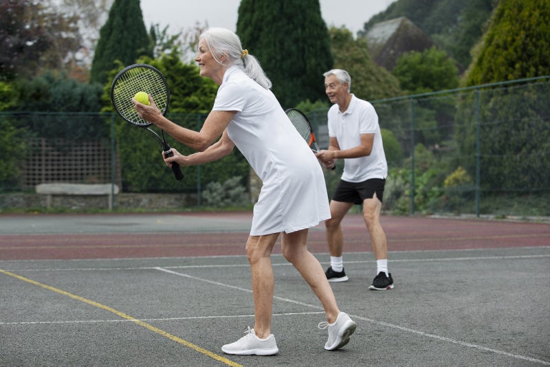 Aging man and woman in the ready stance on a tennis court as the woman prepares to serve the ball.