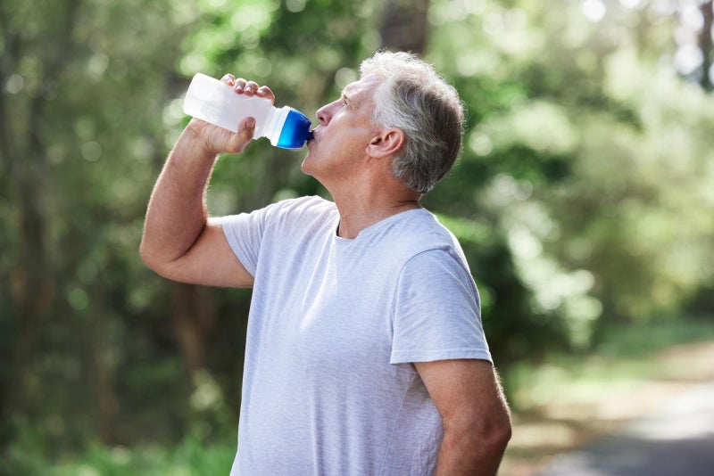 Aging male standing on a wooded pathway taking a swig of clear liquid from a water bottle.