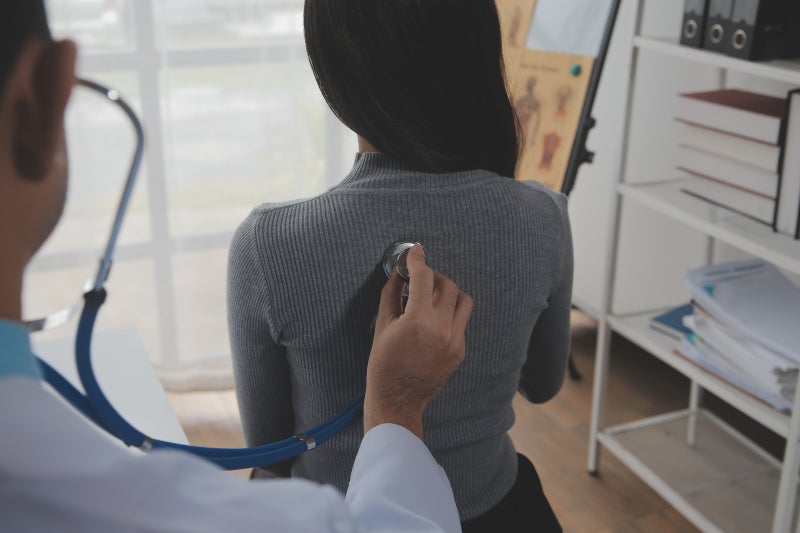 A male healthcare provider in the foreground holds a stethoscope on the back of a dark-haired female seated and facing away from the provider.