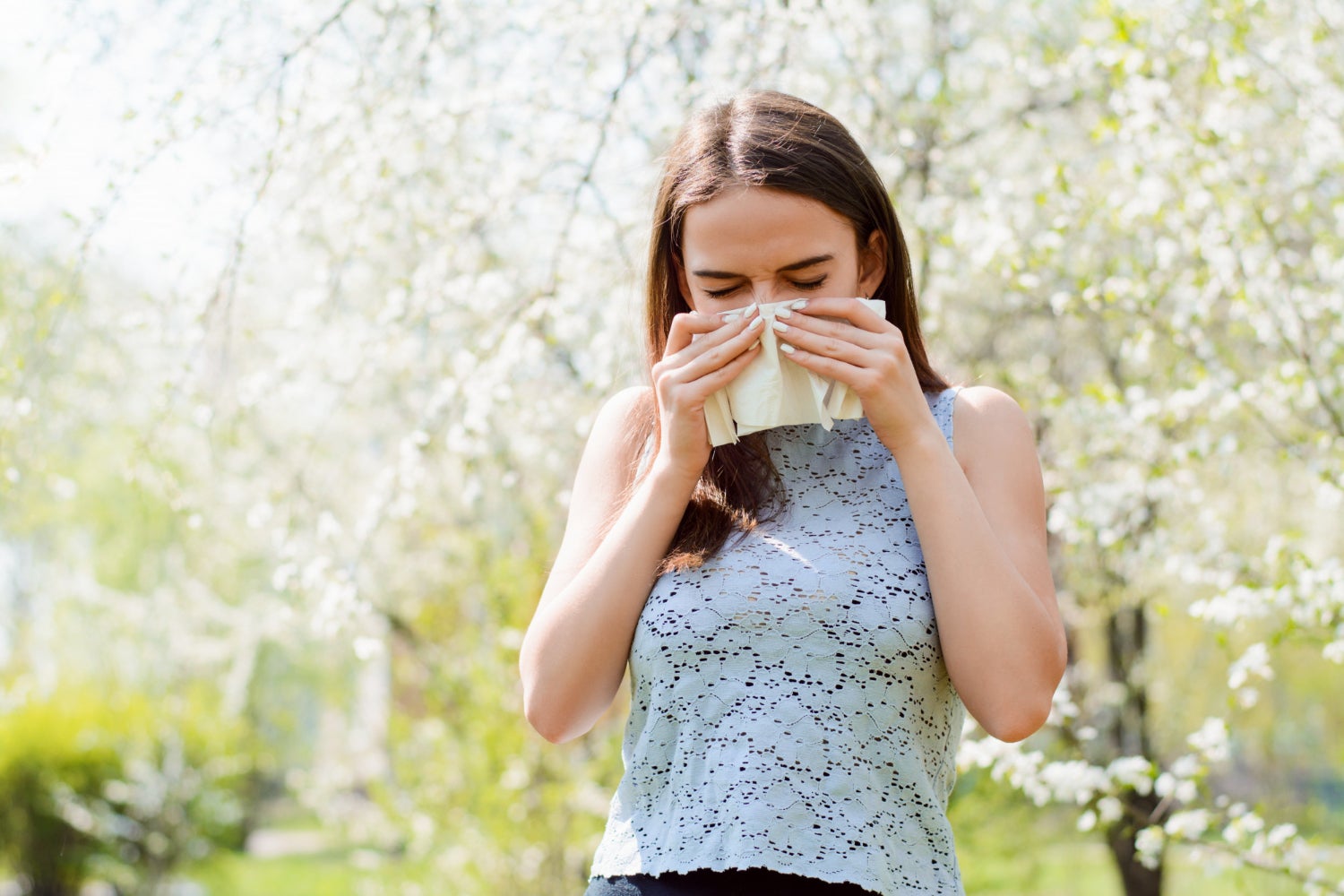 Brunette woman holds a tissue against her nose as she stands outside in front of blooming trees.