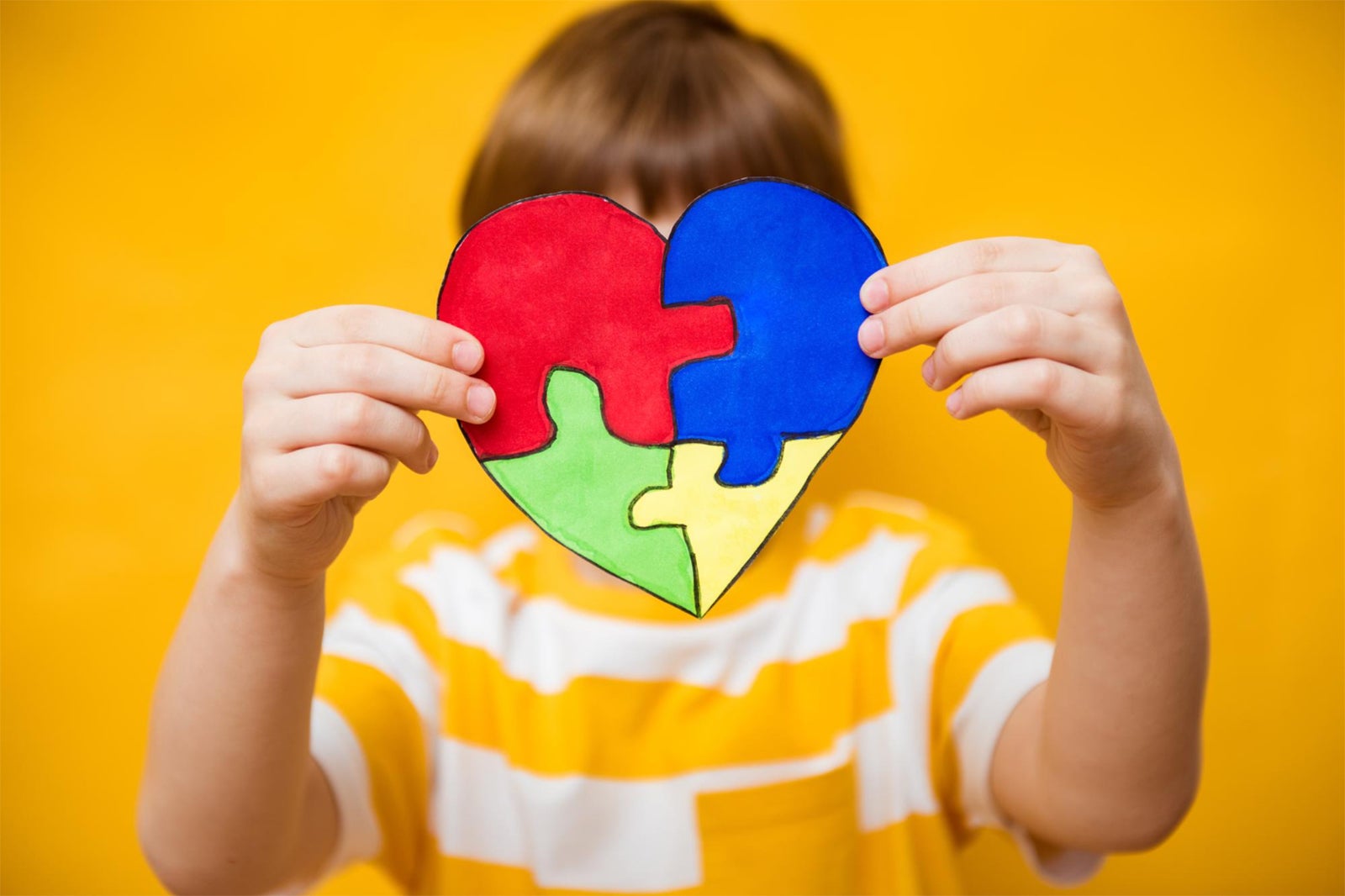 Young male child holding together a heart-shaped, multi-colored four-piece puzzle in front of his face.