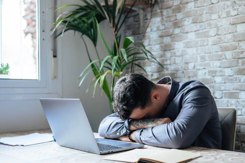 Young Caucasian male sits at a desk with his head down on his arms sitting in front of a laptop.
