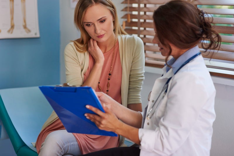Female patient sits on an exam table looking at a clipboard that a female medical provider is showing her.