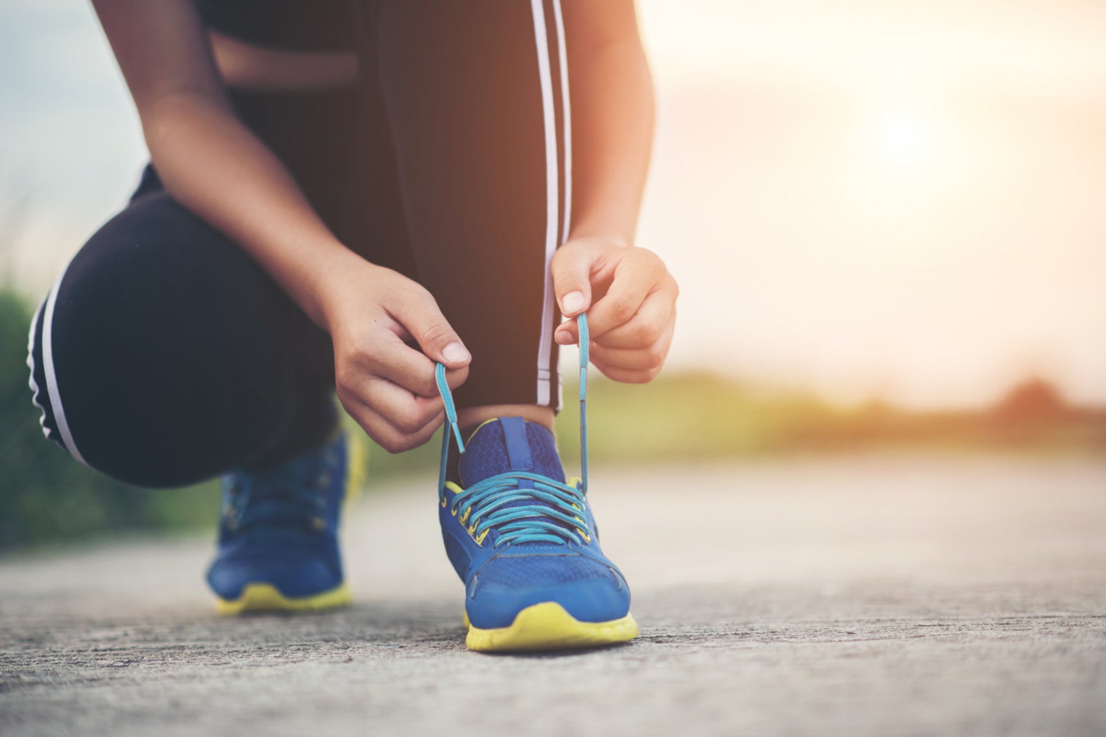 Female runner bends down to tie the laces of a blue sneaker on her left foot on pavement.