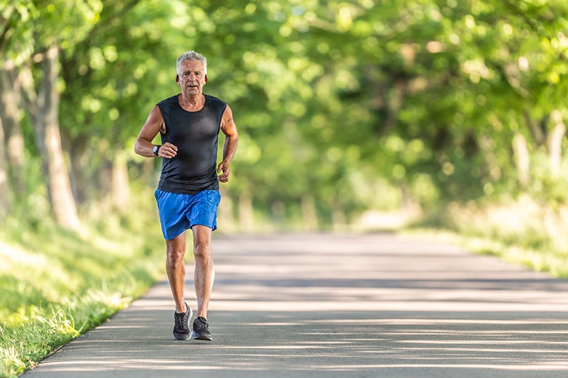 Middle-aged man running along a paved, tree-lined pathway, wearing a tank top and shorts.
