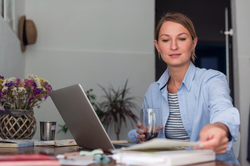 A middle-aged woman sits at a desk in a home setting with an open laptop and a book in front of her as she holds a mug of coffee.
