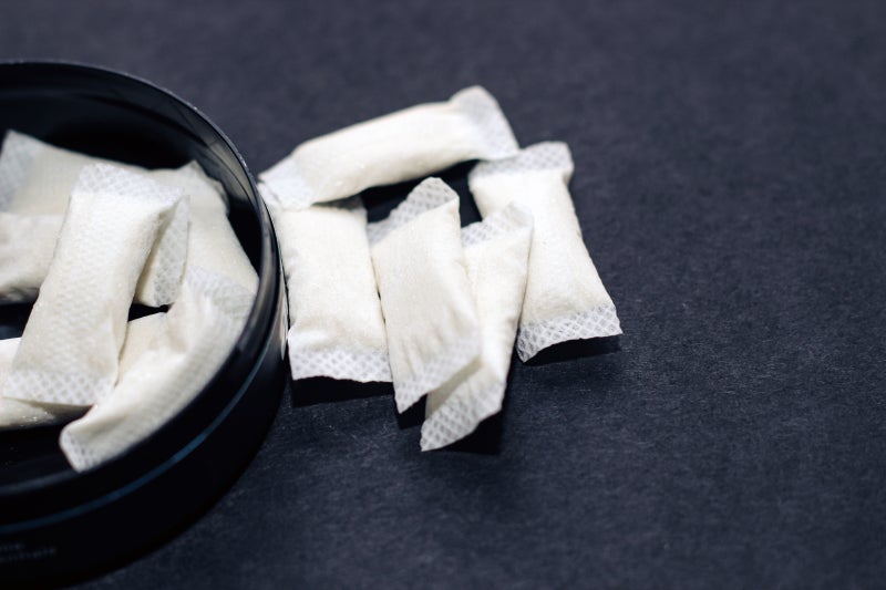 White nicotine pouches lay on a flat, dark surface, outside a container with additional pouches.