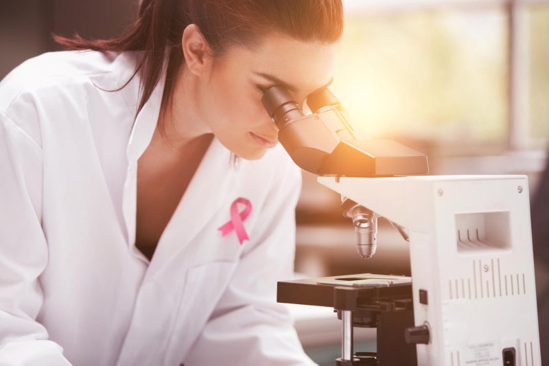 Female scientist wearing a white lab coat with a pink cancer ribbon on the left chest panel leans down to look through a microscope.