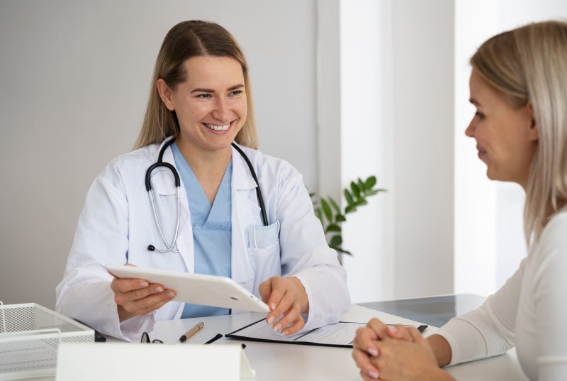 A female healthcare provider smiles at a female patient while both sit at a table or desk.