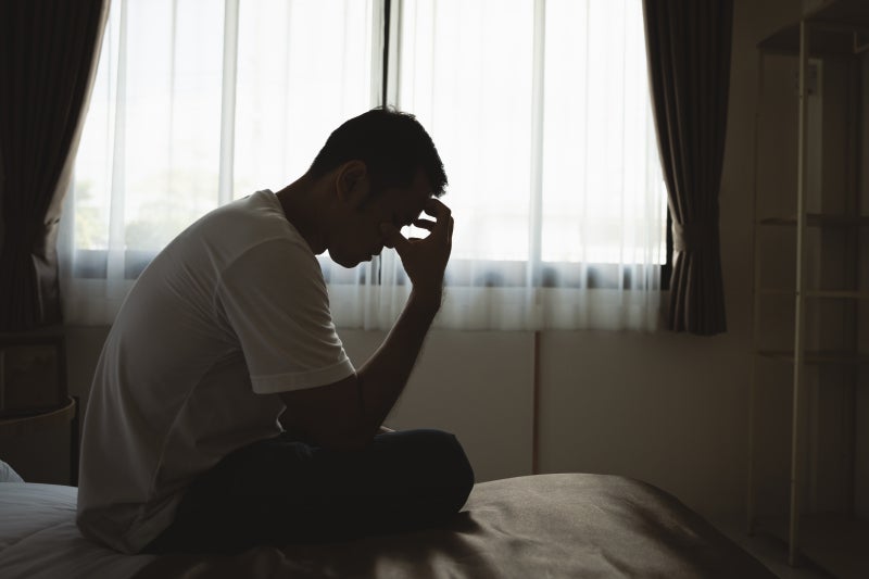 Silhouette of a depressed man sadly sitting on a bed in a bedroom.
