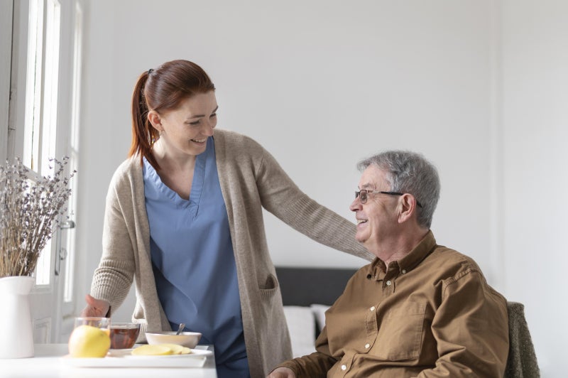 A female healthcare worker stands next to a seated elderly man who is seated at table, both smiling at each other.