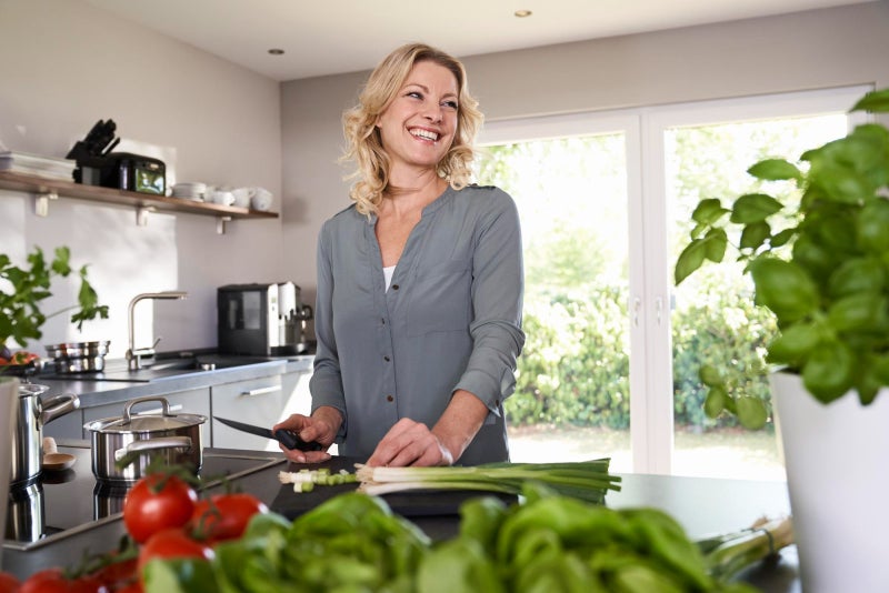 Smiling mature woman standing at a kitchen counter cutting vegetables.