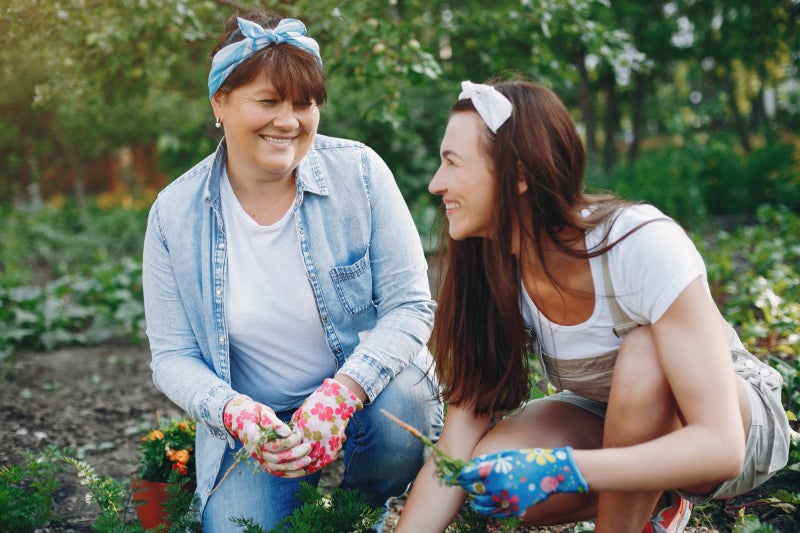 Two dark-haired adult women crouch down on a knee in a garden setting, each wearing a pair of gardening gloves and smiling at each other.