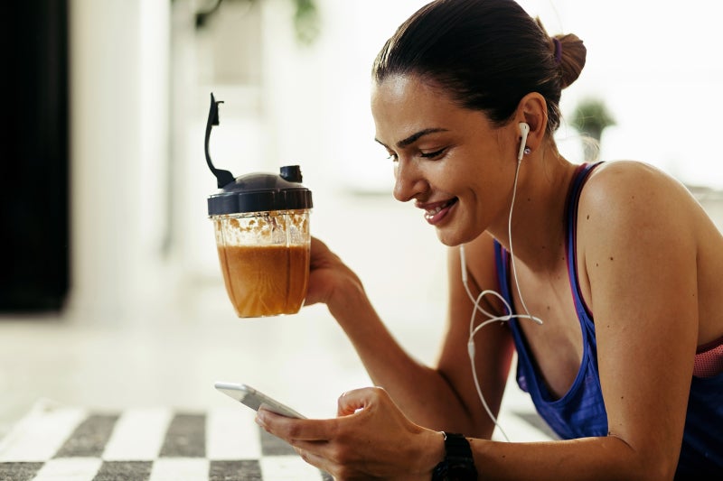 Woman looking at her phone in her left hand while holding a nutrition shake in her right hand with earbuds in her ears.