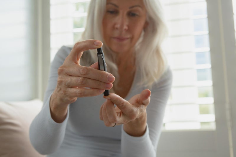 Woman wearing light grey shirt holding left hand out and pricking forefinger to test blood sugar levels