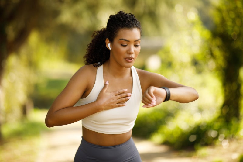 Woman running on a wooded pathway looking at a watch on her left hand and holding her right hand to her chest.