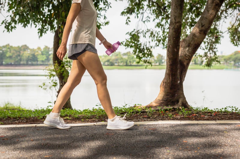 Unidentifiable woman wearing an athletic t-shirt and shorts walking in a park next to a pond with a water bottle in her left hand.