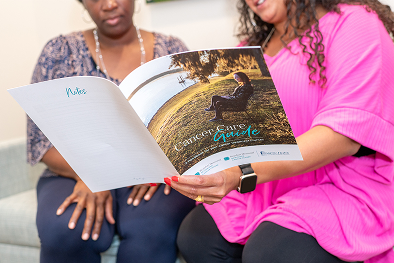 Two unidentifiable women sit on a couch. One is holding open a Beaufort Memorial Cancer Care Guide while the other looks on
